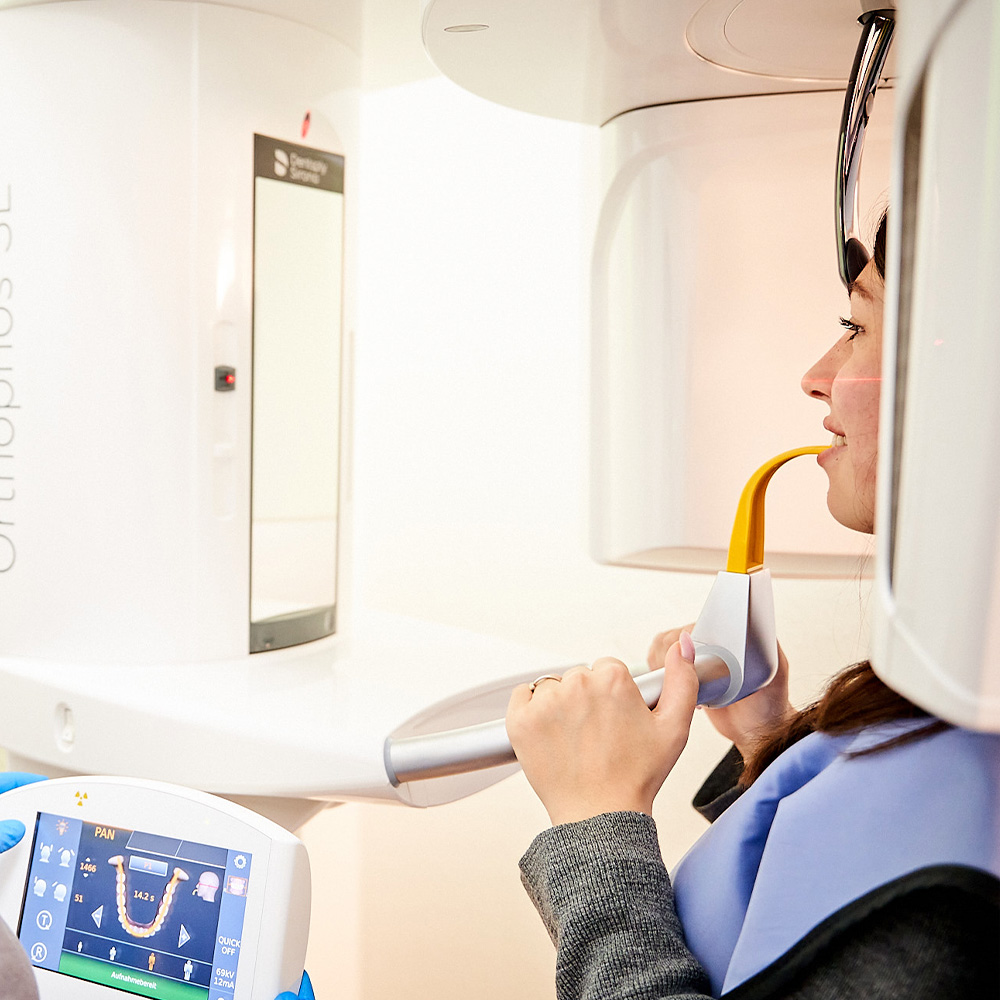 Symbolbild für die Implantologie-Vorbereitung: Patientin steht aufrecht im DVT-Röntgengerät, sie beißt auf einen Haken, damit sie fixiert steht.