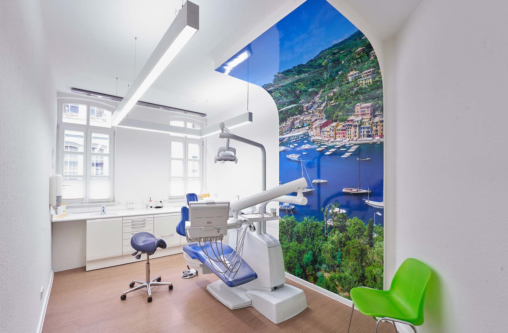 Blick in den Behandlungsraum der Zahnarztpraxis Holz in Wetzlar. Er ist ganz weiß gehalten und ein überdimensionales Bild eines vermutlich italienischen Orts, welches sich hochkant bis an die Decke erstreckt.
